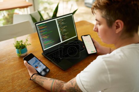 Concentrated individual with tattoos uses tech for productivity, managing tasks in casual setting. Person at wooden table multitasks with coding on laptop, analyzes data on two smartphones.