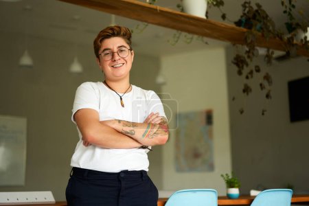 Transgender young adult smiles, arms crossed, wearing casual attire, representing workplace inclusion, gender identity pride. Confident Gen Z individual with tattoos stands in modern office.