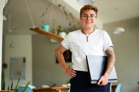 Confident stance inclusive work culture advocate. Transgender professional in bright office smiles, holding laptop, inked arms visible. Nerdy glasses smart casual work attire. Plants modern workspace.