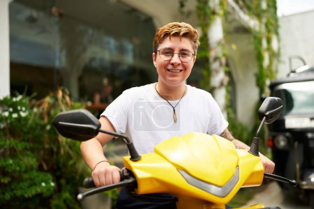 Shirt, shorts, glasses on sunny day, possibly prepping for ride working gig., e-scooter, eco-friendly trend. Smiling transgender person stands with yellow scooter in tropical setting, looks at camera.