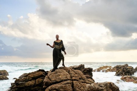 Gender fluid black person poses gracefully standing on rocks in the ocean . Androgynous ethnic fashion model in posh dress and jewellery on rocky beach by storm. Pride month