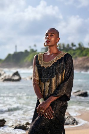 Outrageous gay black person in a luxury gown poses on scenic ocean beach. Non-binary ethnic fashion model in posh outfit with makeup stands gracefully at picturesque location with sea, lighthouse.