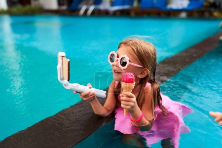 Wearing pink swimwear, heart-shaped sunglasses, she smiles for followers. Little girl enjoys ice cream, streams live in pool. Casual summer scene, child influencer shares moments, cool water backdrop.