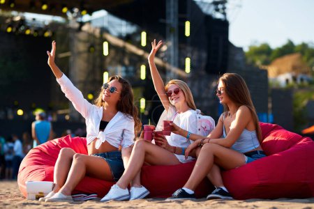 Friends in casual wear smile, wave hands, celebrate at seaside concert event. Group of happy girls toast drinks on red bean bag chairs, enjoy summer music festival on sandy beach, cheer in sunlight.