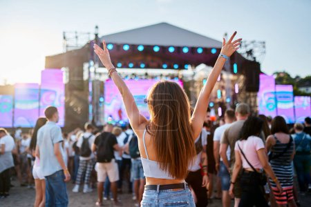 Friends dance, celebrate in crowd, summer vibe at coastal event. Back view of girl with raised arms, enjoying live music at sunset beach festival. Teens in casual wear, fun at sunny seaside gig.