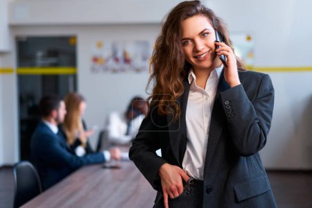 Confident caucasian blonde businesswoman in smart casual attire talks on mobile phone in a modern office standing with colleagues in background having a meeting.