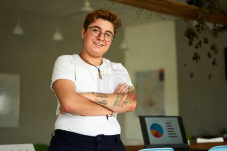 Inclusive work environment, laptop with charts in background. Confident transgender professional stands in modern office, arms crossed, smiling. Pride, equality themes in corporate setting.