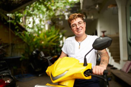 Person ready for ride, embracing freedom, warm climate. Smiling transgender individual on yellow scooter enjoys tropical setting. Confident pose with bike, plant backdrop signals leisure, mobility.
