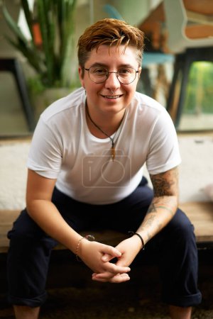 Tattooed, stylish glasses, modern diversity, gender fluidity. Smiling person sits in vibrant coworking space, reflecting Gen Z vibe. Warm ambient enhances welcoming, inclusive work environment.