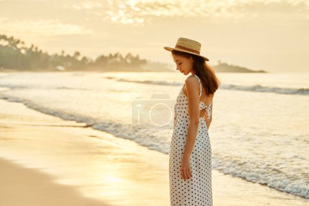 Entspanntes Tempo, Meeresbrise, goldenes Stundenlicht, verkörpert Freizeit, Urlaubsstimmung in der Nähe von Meereswellen. Elegante Frau spaziert im Sommer am sonnigen Strand, weißes gepunktetes Kleid, Strohhut auf.