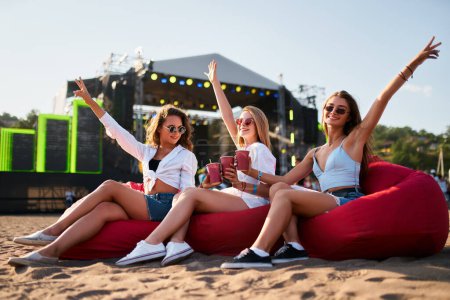 Téléchargez les photos : Trois femmes se détendent sur des chaises de sac de haricot rouge au festival de musique de plage, journée ensoleillée, élever des boissons, scène en arrière-plan, profiter des vibrations d'été, lunettes de soleil, vêtements décontractés, bras levés gai concert amusant. - en image libre de droit