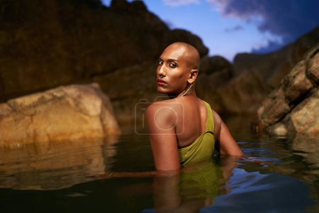 Androgynous black fashion model poses chest deep inside natural pool surrouded by rocks on tropical island at night. Trans sexual ethnic fashion model in the middle of backwater exotic scenic location