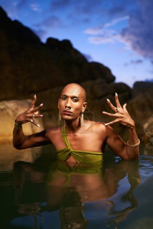Photo for Gender fluid black person poses in natural still water pool. Queer ethnic fashion model in long revealing clothes, brass jewelry with gems standing gracefully in the middle of a rocky lake at night. - Royalty Free Image