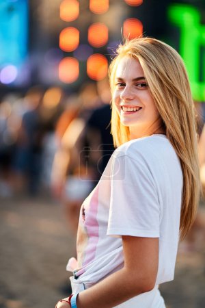 Crowd gathers behind for live concert experience. Blonde woman smiles at sunset, enjoying outdoor music event on beach. Summer, carefree youth lifestyle in focus. Casual fashion, fun atmosphere.