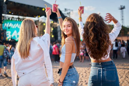 Summer event scene with vibrant energy, youth lifestyle, friendship. Back view three girls enjoy music fest on sandy beach, hold colorful drinks, dance near stage. Sunset lights casual outdoor party.