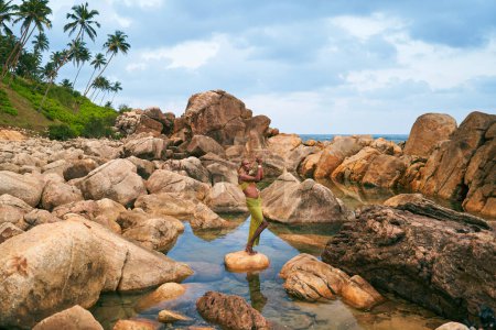 Transsexuelles schwarzes Modemodel posiert in einem natürlichen Pool, der von Felsen auf einer tropischen Insel umgeben ist. Androgyne ethnische Mode-Modell steht auf Stein in der Mitte von Rückstau an exotischer szenischer Lage