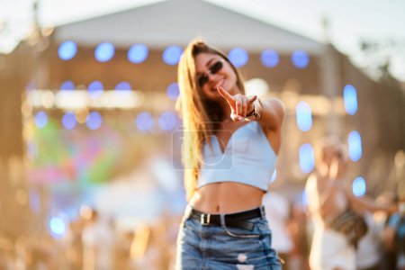 Casual outfit, sunlight, crowd. Happy young woman dances at beach music fest, summer vibe. Lady enjoys live concert, points, smiles. Youth lifestyle, entertainment begins at outdoor event.