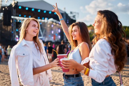 Smiling females in casual summer attire enjoy live concert with friends on sandy shore. Group of young women cheering with drinks at beach music festival, sunlit stage in background.
