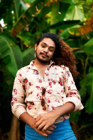 Flamboyant pose, bright floral shirt open, denim pants. Confident gay man with curly hair stands in tropical setting. Arms crossed, smiling, gender expression, lush green foliage backdrop.