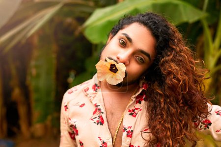 Retrato exótico, trasfondo de la naturaleza, orgullo LGBTQ. Hombre del sur de Asia con flor en la boca expresa con confianza la feminidad en el entorno tropical. Cabello rizado, maquillaje audaz, aspecto elegante, atuendo vibrante.