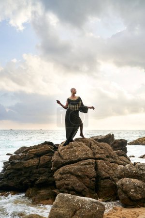 Gender fluid black person poses gracefully standing on rocks in the ocean . Androgynous ethnic fashion model in posh dress and jewellery on rocky beach by storm. Pride month