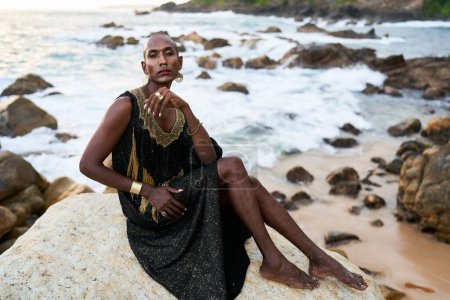 Modelo de moda bipoc no binario en vestido, joyas de latón se sienta en las rocas por el océano. Trans persona negra sexual con anillos, anillo en la nariz, pulseras, pendientes en ropa elegante posa en lugar tropical junto al mar.