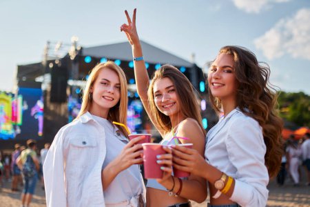 Girls celebrate with drinks, smile with stage backdrop, sunny seaside vibes. Group of young women clink cups at beachside music event. Partygoers enjoy festival atmosphere, blue sky, summer leisure.