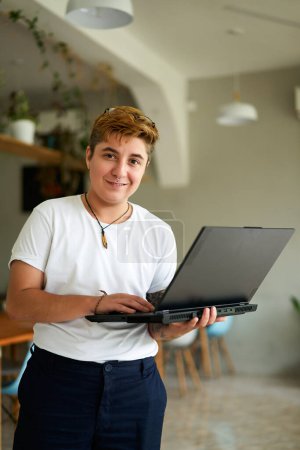 Short-haired, smiling FTM pro in casual wear. Confident transgender employee holds laptop in modern office cafe. Inclusive workplace, gender diversity, tech job.