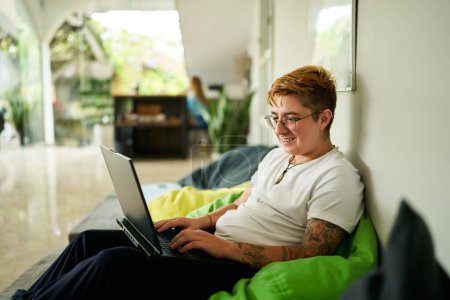 Transgender person lounges on bean bag chair, working on laptop in a light-filled coworking space reflecting modern work flexibility and inclusion in the professional environment.