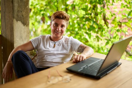 Inclusive remote work scene captures gender diversity, modern freelancing trend amongst rich plants. Transgender Gen Z with tattoos smiles, works on laptop in tropical coworking space.
