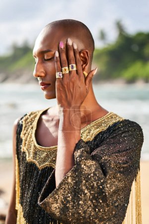 Outrageous gay black man in luxury gown, jewelry poses on scenic ocean beach. Gender fluid ethnic fashion model in a posh dress, accessories looks at camera covers face with hand and rings on fingers.