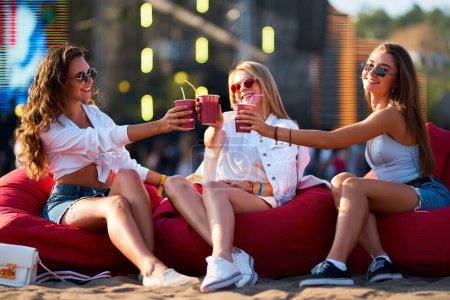 Trois amis insouciants en tenue d'été, des lunettes de soleil clink tasses, se détendre sur la côte. Groupe de jeunes femmes heureuses assises sur des sacs de haricots rouges au festival de musique balnéaire ensoleillée, acclamations avec boissons, vibrations fraîches.