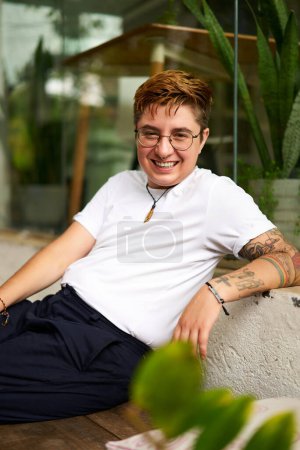 Gen Z Individual with tattoos break exudes confidence, inclusion atmosphere. Smiling transgender person sits casually, modern coworking space with plants in background. Casual smart creative office.