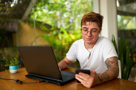 Casual business setting, gender diversity at work. Transgender office worker with tattoos checks smartphone, distracted from laptop. Indoor plant, natural light, inclusion in modern workplace.