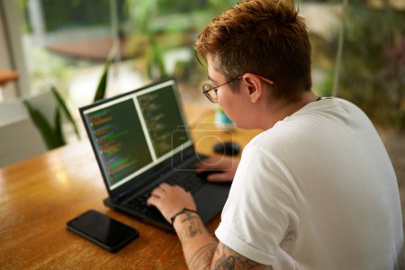 Focused individual with tattoos types code, designs app. Transgender web developer works on laptop, programming in modern eco office. Commitment to inclusivity, equality in tech industry workplace.