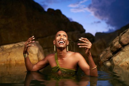 Lächelndes bipoc lgbtq Modell posiert nachts im Wasser innerhalb eines natürlichen Pools. Nichtbinäre Person zeigt Schmuck - Ringe mit Edelsteinen an den Fingern, Messingnasenring, goldene Ohrringe, Armbänder, Ständer in einem Teich.