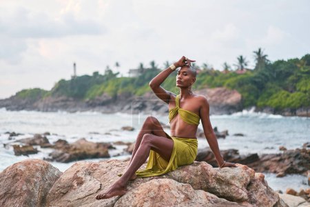 Non-binary black person poses gracefully sitting on top of rock in ocean at sunset. Trans ethnic divine fashion model poses in open tropical dress on peak by a storm at dusk against sunrise. LGBT