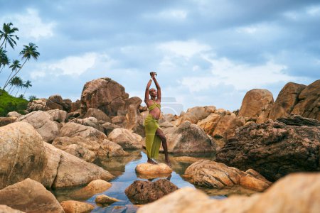 Una persona negra no binaria está parada sobre una roca en medio de agua quieta dentro de un arroyo. Lgbtq modelo de moda elegante étnica posa de pie sobre una piedra en un estanque cristalino en un lugar tropical exótico.