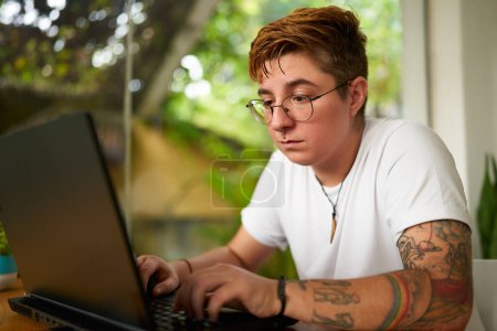 Gender diversity in remote work setup, inclusive workplace ambiance with home plants. Transgender male concentrates on freelance work from home, using laptop, glasses on, tattoo visible.