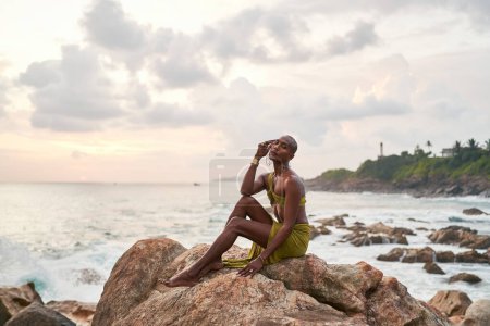 Homosexuell bipoc anmutiger Mann posiert hoch oben auf Felsen im Ozean bei Sonnenuntergang. Homosexuelle schlanke ethnische Modemodel in einem tropischen Maxi-Kleid auf einem felsigen Hügel über Sturm in der Dämmerung. Stolz auf LGBTQIA