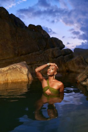 Gender fluid black person dives out from the water in a natural still water pool. Queer ethnic fashion model in long revealing clothes poses gracefully waist deep in middle of clear lake at night.