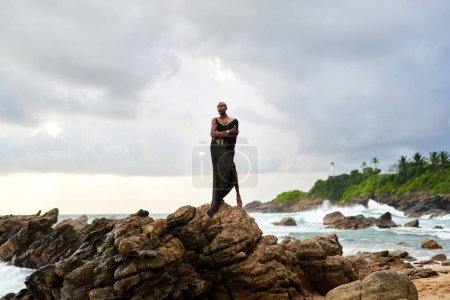 Non-binary black godlike person poses gracefully standing on rocks in ocean. Trans ethnic fashion model in a posh dress and jewellery on rocky beach by storm . Lgbtq. Pride month