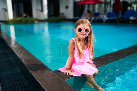 Smiles on sunny day, feet dipping in water. Little girl sits by poolside in pink dress, sunglasses. Kid enjoys luxury resort, summer vibes. Fun childhood moment, family vacation concept.