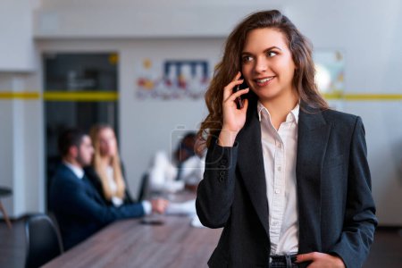 Female employee communicates on mobile during work, team discusses project behind. Confident caucasian blonde businesswoman talks on smartphone in modern office, standing with hand in pocket.