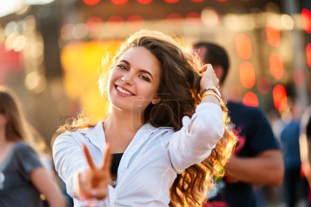 Photo for Casual outfit, dancing on sand, joyous mood, crowd blur in background, reaching out to camera for an inviting vibe. Happy young woman enjoys summer music fest on beach with sunset backlighting. - Royalty Free Image