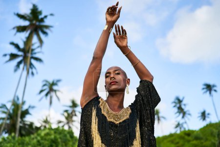 Modèle de mode noir androgène se dresse sur la plage pittoresque de palmiers tropicaux portrait. Une personne ethnique non binaire portant du maquillage, des bijoux pose les mains en l'air à un endroit exotique. Trans sexuelle. Mois de fierté.