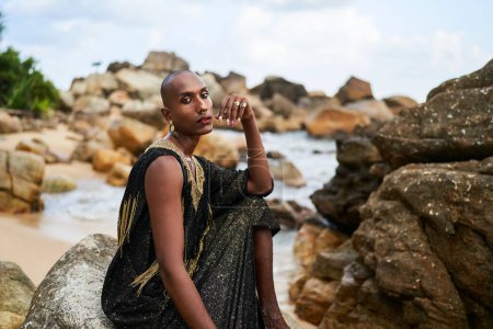 Non-binary ethnic fashion model in a luxury dress sits on rocks by the ocean. Trans sexual black person in jewellery and posh clothes poses gracefully in tropical seaside location.