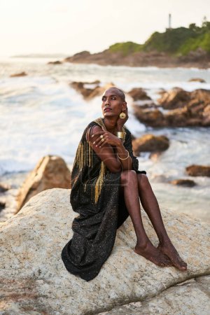 Nicht-binäre schwarze Person im Luxuskleid, Goldschmuck am Strand Felsen im Ozean. Transethnisches Mode-Model mit Schmuck im schicken Kleid posiert anmutig in tropischer Küstenlage bei Sonnenuntergang.