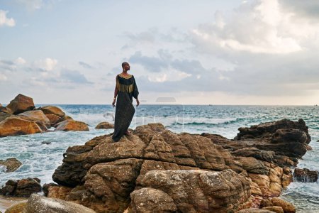 Photo for Non-binary black godlike person poses gracefully standing on rocks in ocean. Trans ethnic fashion model in a posh dress and jewellery on rocky beach by storm . Lgbtq. Pride month - Royalty Free Image