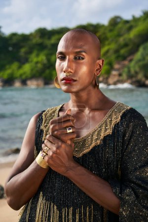 Gorgeous gay black man in luxury gown, jewelry poses on scenic ocean beach . Non-binary ethnic fashion model wears long posh dress golden rings, earrings, bracelets, looks at a camera.